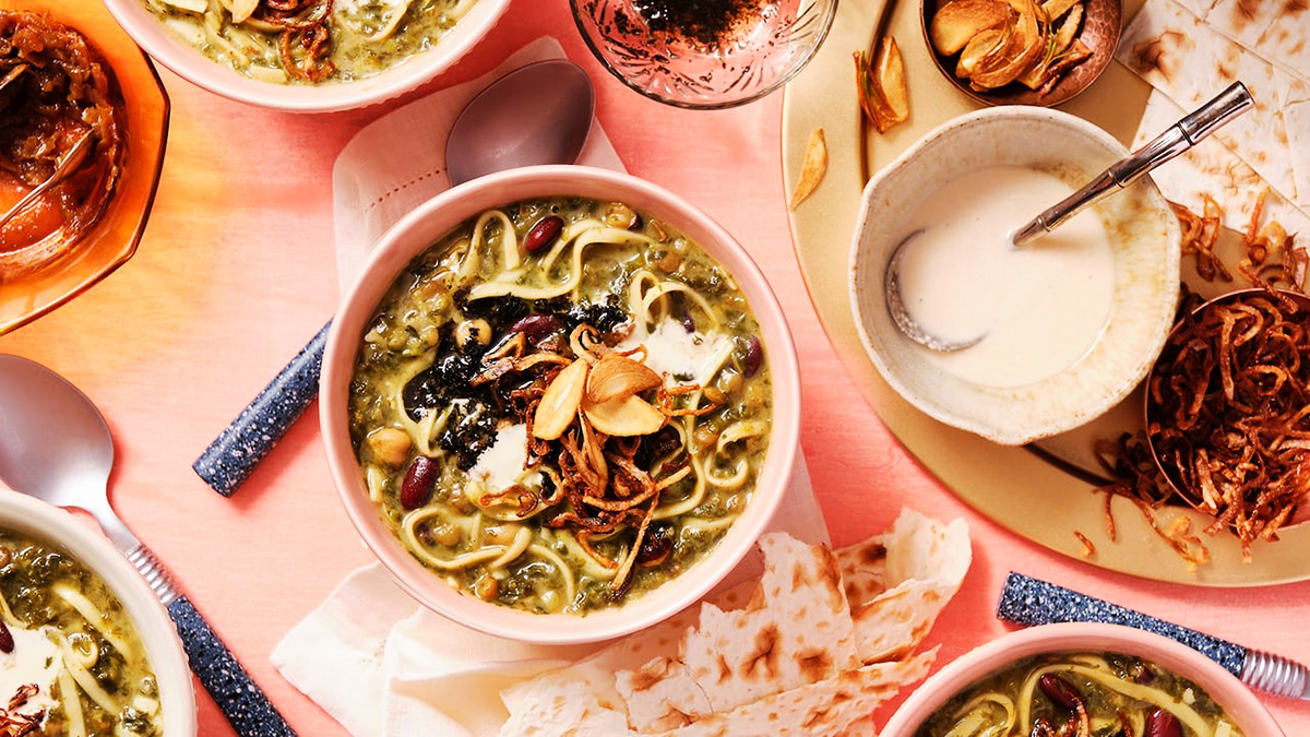 تصویر یک میز که شامل یک چند کاسه آش رشته تزئین شده با نعنا داغ و کشک و پیاز و سیر داغ، کشک، قاشق با نقش و نگار ایرانی و پیاز داغ است